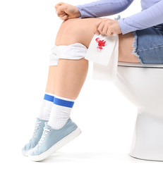 Young woman with paper in blood sitting on toilet bowl against white background. Hemorrhoids concept