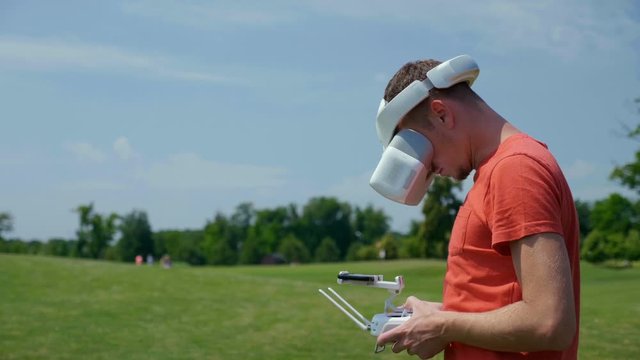 Man controls a quadrocopter through a remote control and looks at the video with goggles on his head. Drone operator in a red T-shirt and blue shorts in park is filming drone video