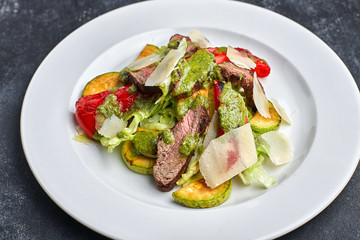 Warm salad with veal, baked vegetables, bell pepper, zucchini and parrmesan cheese, on a white plate, against a dark background