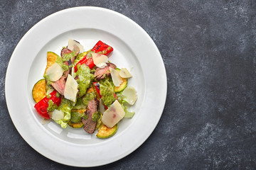 Warm salad with veal, baked vegetables, bell pepper, zucchini and parrmesan cheese, on a white plate, against a dark background