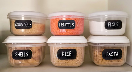 Six plastic containers with dry food (couscous, red lentils, white flour, pasta shells, rice, elbow pasta) in the kitchen cupboard.