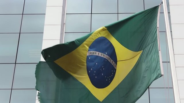 Storm winds wildly blowing a large frayed Brazilian flag on flagpole against a modern high rise corporate window building in the background. 60fps normal speed