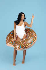 Fit woman dressed in swimsuit posing happy with donut inflatable showing peace gesture circle over blue background.