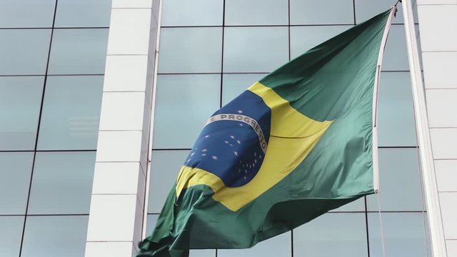 Strong winds blowing a large frayed Brazilian flag on flagpole with wild dramatic texture and folds against a modern high rise corporate window building in the background. Slowed down from 60fps.