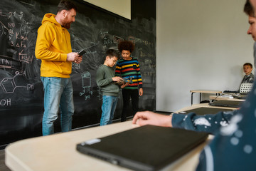 Fototapeta na wymiar Center For Excellence. Young male science teacher standing near the blackboard, holding tablet pc and looking at his students while they are demonstrating their own robot vehicle