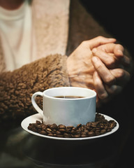 Manos de mujer adulta calentandose al costado de una taza de cafe caliente con granos tostados alrededor sobre un plato blanco redondo y fondo negro.