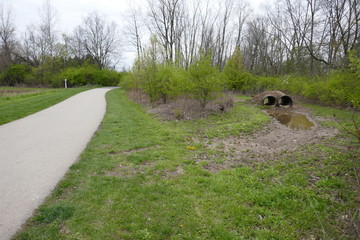 Paved walking path through public park with drainage pipes and water overflow to side
