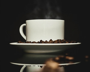 Toma lateral de taza de café caliente rodeada de granos de cafe tostado sobre un plato redondo blanco en fondo negro con reflejo.