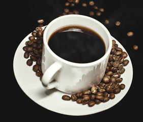 Toma lateral superior de taza de café caliente rodeada de granos de cafe tostado sobre un plato redondo blanco en fondo negro.