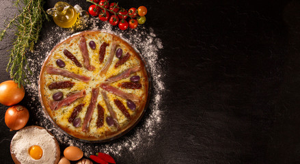 Tasty prosciutto pizza and cooking ingredients tomatoes basil on black concrete background. Top view prosciutto pizza