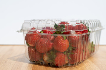 Fresh red organic strawberries in a clear plastic grocery container. The strawberries have green leaves on the berries. The plastic package is sitting on a wooden table with a white background. 