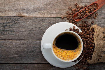 Top view of Hot fresh black coffee for morning with milk foam. Breakfast menu in white ceramic cup with coffee beans roasted in burlap sack bag on wood table background. Flat lay with copy space.