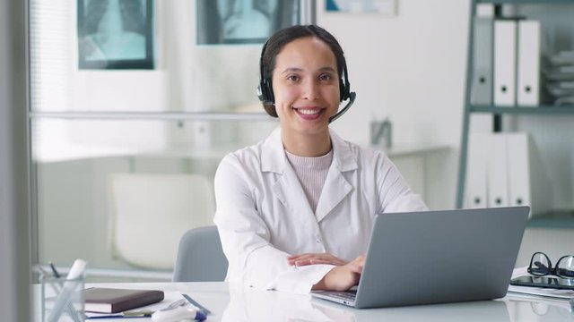 Positive young female doctor in lab coat and wireless headset sitting at desk with laptop on it, looking at camera and smiling during workday in medical office