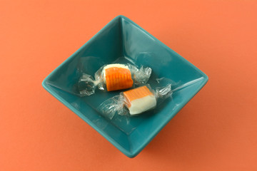 Two orange and champagne flavored chew candy pieces in plastic wrappers in blue square bowl on orange background