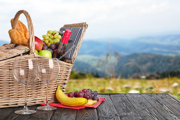 Wicker picnic basket with wine and different products on wooden table in park, space for text