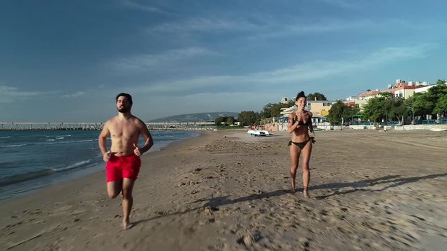 Young Caucasian people wearing bathing suits jogging on sandy beach, aerial between people, slow motion