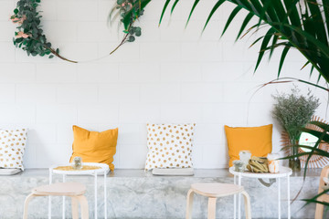 A modern decoration and relaxing cement seat with white wall,  yellow and dotted cushions, grey seat cushion, flowers, 2 drinks on the white coffee table, soft focus tree as a photo frame.