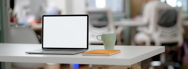 Cropped shot of simple office desk with blank screen laptop, mug and notebook