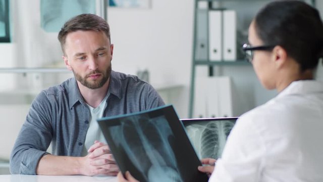 Handsome Caucasian man sitting at table in medical office, looking at x-ray scan of lunges and discussing diagnosis with female physician during consultation