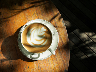 Latte art coffee on a plate with a white pattern Set on a wooden table in a coffee shop The sunlight in the evening shines through the trees, causing shadows on the coffee mugs.