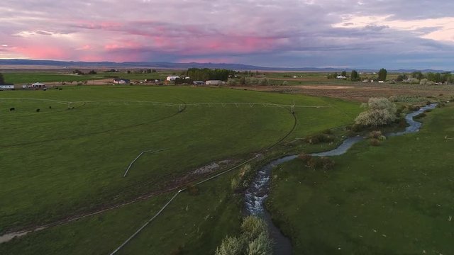 Aerial view over farmland in Idaho during colorful sunset moving along small river canal through farm fields and pastures.