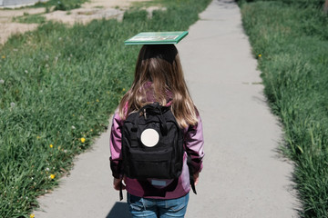 little girl going to school with schoolbag and a book on a head top. girl with backpack outdoors...