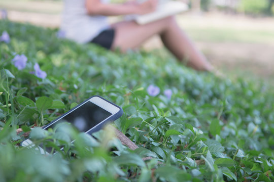 Digital detox concept. Lonely smartphone on grass because Asian beautiful woman reading a book for relaxing activity without digital technology in the park on natural light freshness morning