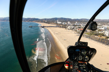 Helicopter cockpit flying on landscape and blue sky, with pilot arm driving in cabin. Spectacular view of ocean, curve road, mountains, Santa Monica Pier