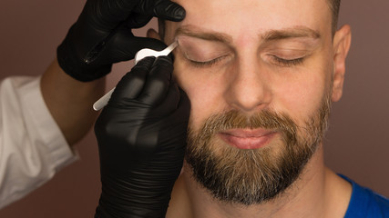 Professional barber doing threading procedure and correcting shape of eyebrows to young male client with tweezer in barber shop. Barber at work. Brow care concept. Plucking brows