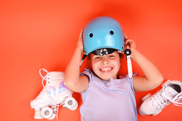 A little child sits with roller skates in a blue helmet with a happy smile and showing a peace sign. A girl of 7 years is preparing for active leisure on skates. Copy space