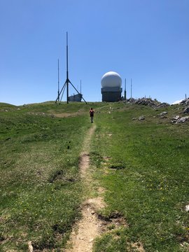 White Radome: Round Surveillance Radar Tower Dome For Air Traffic And Meteorology, Communication Antennas In Nature, Blue Sky. Woman Hiking On A Path In The Grass Towards The Modern Installation.