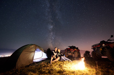 Man and woman sitting together near tourist tent, burning campfire, two atv quad motorbikes on the top of mountain, romantic couple enjoying beautiful view of night sky full of stars and Milky way © anatoliy_gleb