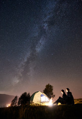 Back view of happy couple travellers sitting at bonfire beside camp and glowing tourist tent under night sky full of stars and Milky way. On background amazing starry sky, mountains and luminous city