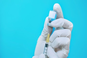 A female Asian physician with white rubber gloves and surgical mask at a clinic, inserting a needle and syringe on a clear glass bottle of vaccine with blank white label to inject it to a patient.