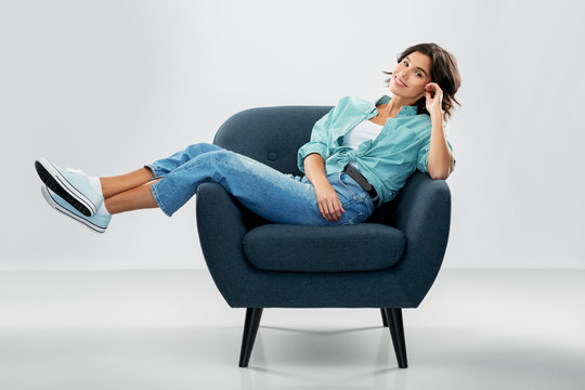 comfort, people and furniture concept - portrait of happy smiling young woman in turquoise shirt and jeans sitting in modern armchair over grey background