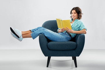 comfort, people and furniture concept - portrait of happy smiling young woman in turquoise shirt and jeans sitting in modern armchair and reading book over grey background