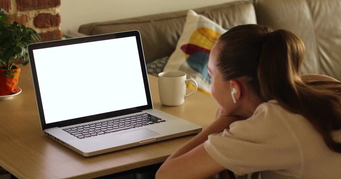 Side view of anonymous female in earbuds listening to music while sitting in front of open netbook with blank screen and mug of hot drink near sofa and brick wall