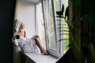 Young beautiful woman in white towel and robe drinking coffee on the windowsill at home.