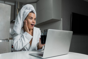 Young beautiful woman in white towel and robe drinking coffee and looking on laptops screen at the table. Morning of a woman at home concept.