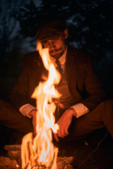 Portrait of a guy sitting by the fire at night in nature. Cinematic vintage photo. Retro 1920s...