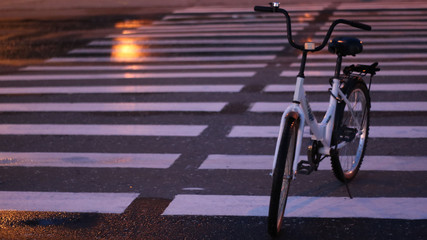 black and white bicycle standing at the pedestrian crossing of an evening wet road on which yellow lights are reflected