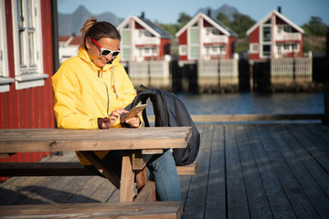 Young woman in a yellow jacket on a background of traditional red Norwegian rorbu houses with a smartphone in hand, traveling to Norway Lofoten Islands