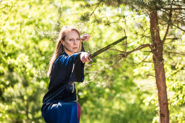 A female warrior with a sword poses on a stump in the forest. Wushu fencing