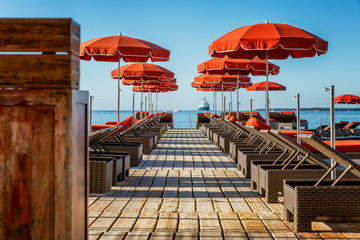 Empty deckchairs with orange mattresses and parasols on wooden flooring on the shore of the blue...