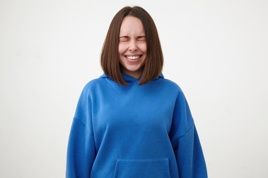 Good looking young pretty short haired brunette female dressed in blue hoodie smiling cheerfully with closed eyes while standing over white background