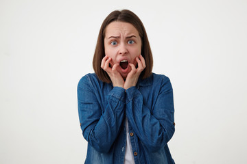 Frightened young short haired brunette woman without makeup holding her face with raised hands and...