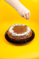 Chocolate cake decorated with flaked coconut, homemade pie on yellow surface. Femake hand scattering coconut flakes