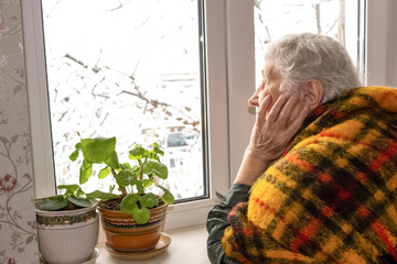 Old lonely woman sitting near the window in his house.