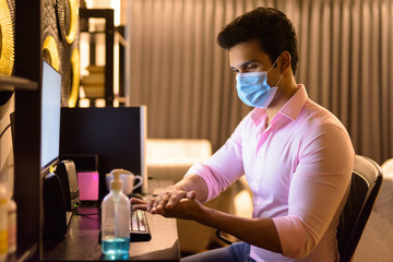 Young Indian businessman with mask using hand sanitizer while working overtime at home during quarantine