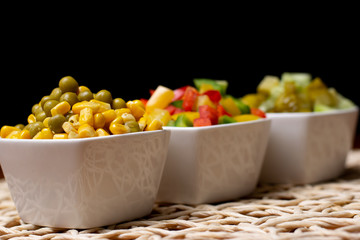 Vegetables in white bowls, diced on a brown kitchen table. Peppers, cucumbers, sweet corn and peas...